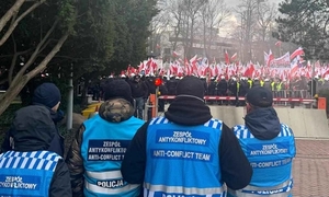 na zdjęciu policjanci w niebieskich kamizelkach z napisem zespół antykonfliktowy stoją przy marszu